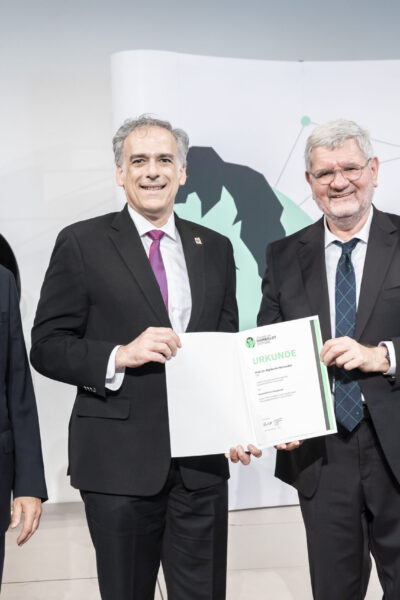 Three men standing, as the one on the left is presenting an award (shown) to the one in the center